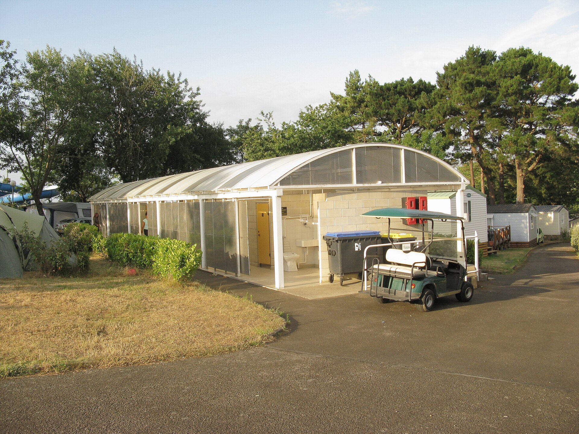 Een open sanitair gebouw op een camping, met een gebogen dak, wasbakken en toiletdeuren, tussen tenten en stacaravans