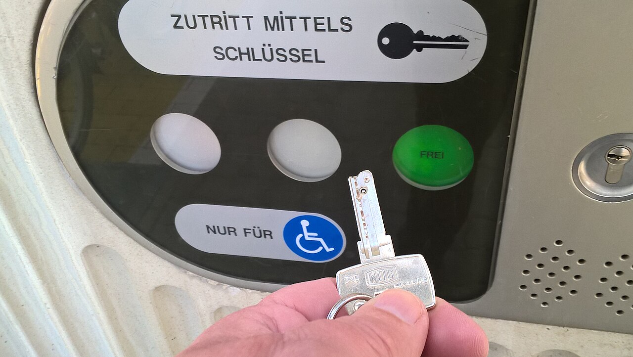 Een hand met een eurosleutel voor het bedieningspaneel van een aangepast toilet, met het opschrift "Zutritt mittels Schlüssel"