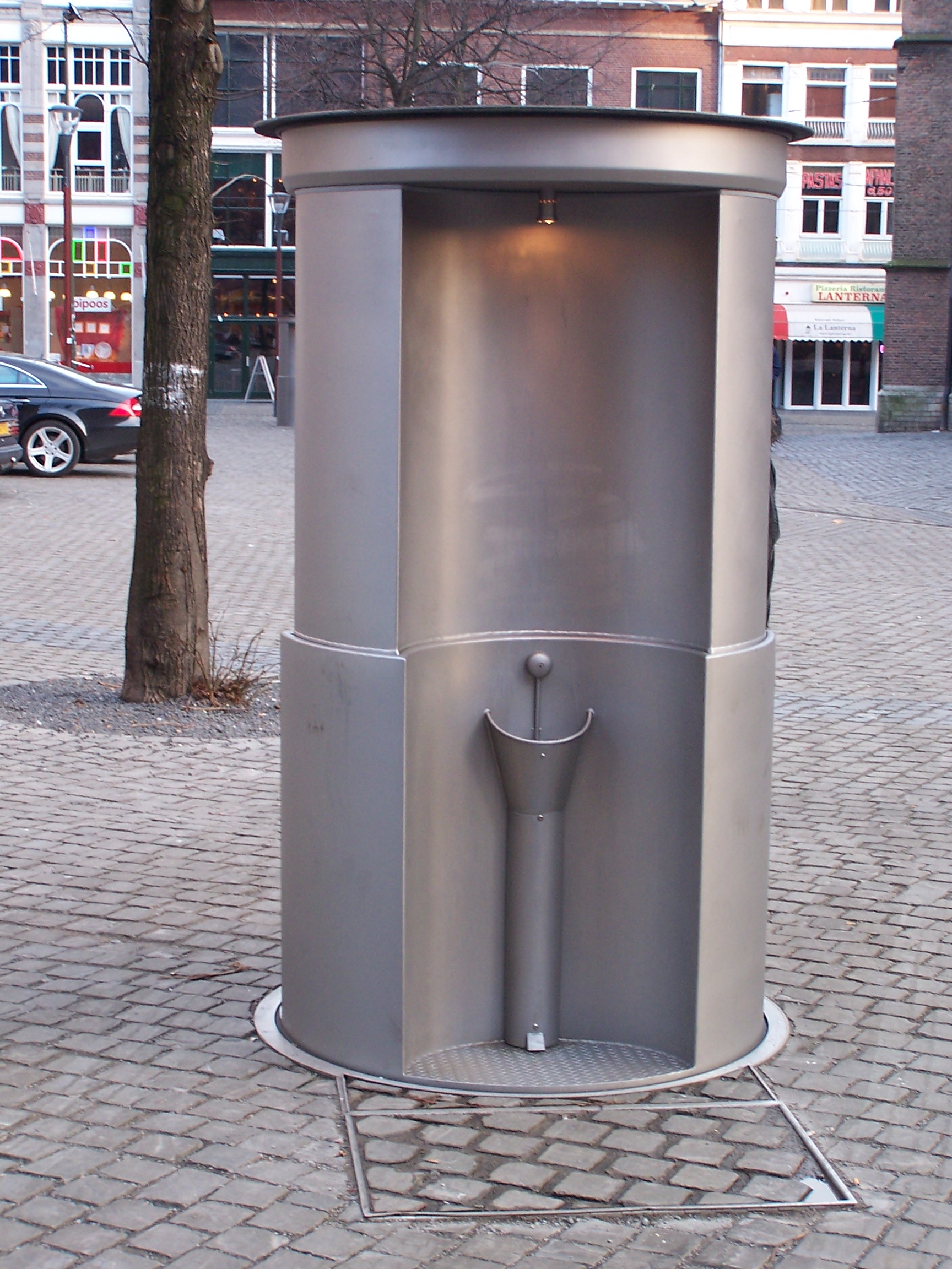 Een opgeheven metalen plaszuil op een plein, met de ronde put in de bestrating waarin hij wegzinkt
