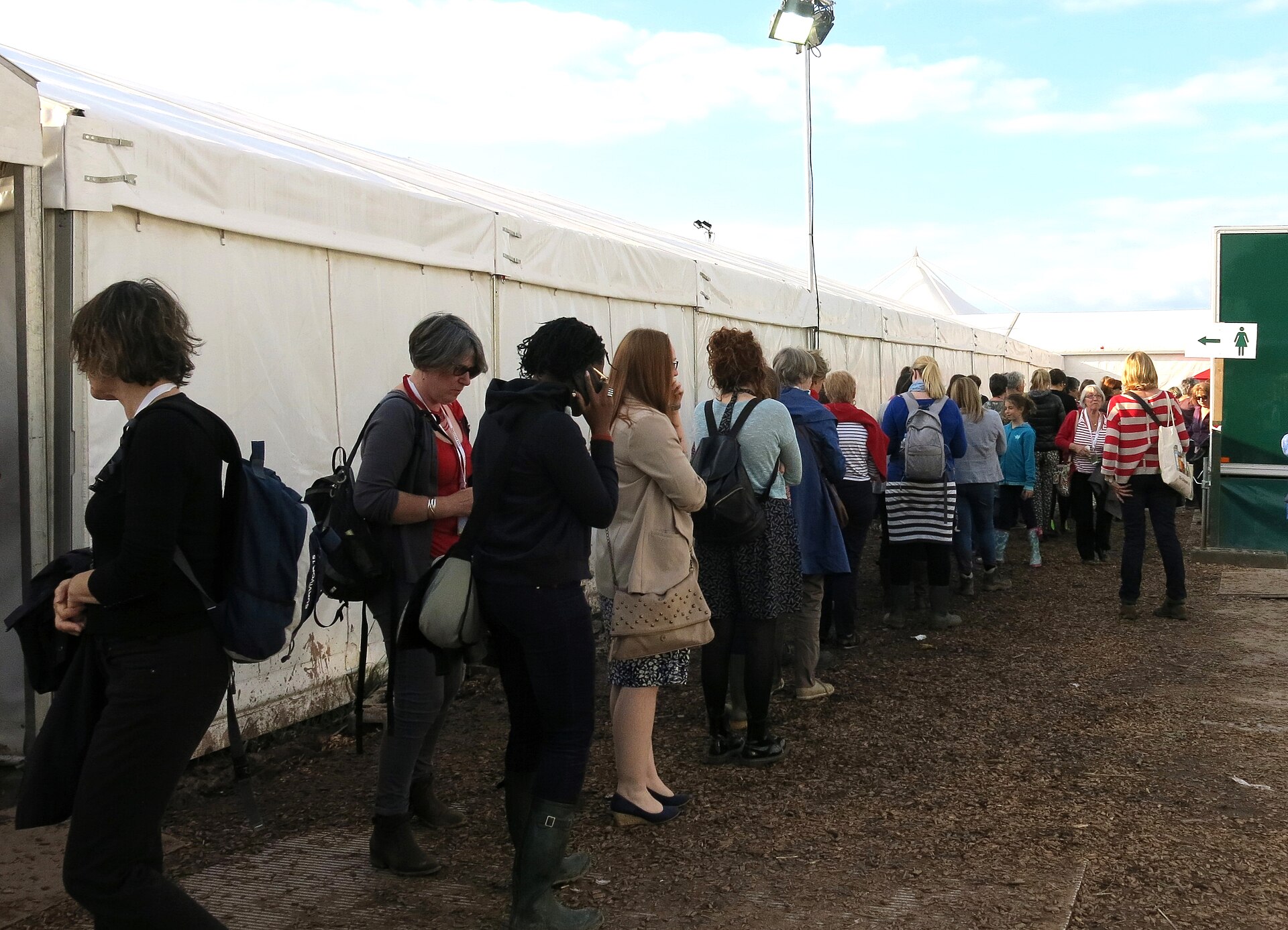 Een lange rij mensen die aanschuiven voor de damestoiletten op een evenemententerrein, met een groen bordje en een pijl